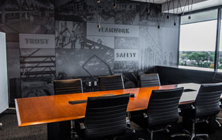 A conference room with a dark gray wall and the words "Trust," "Teamwork," and "Safety" emblazoned on it. A wood conference table flanked by black office chairs sits in the middle of the space.