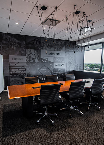 A conference room with a dark gray wall and the words "Trust," "Teamwork," and "Safety" emblazoned on it. A wood conference table flanked by black office chairs sits in the middle of the space.
