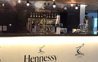 A counter in a bar with the Hennessy logo featured against the wall above the alcohol and under the bar front.