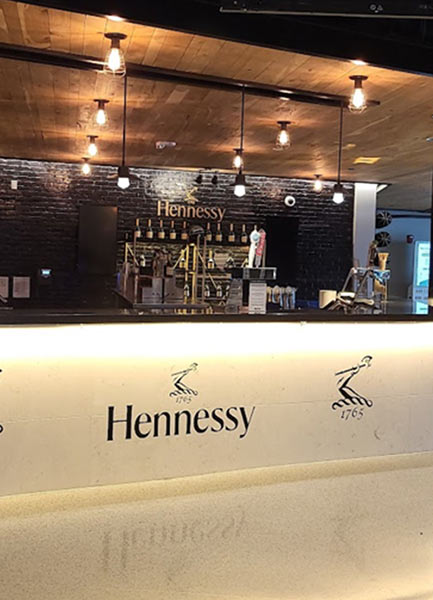 A counter in a bar with the Hennessy logo featured against the wall above the alcohol and under the bar front.