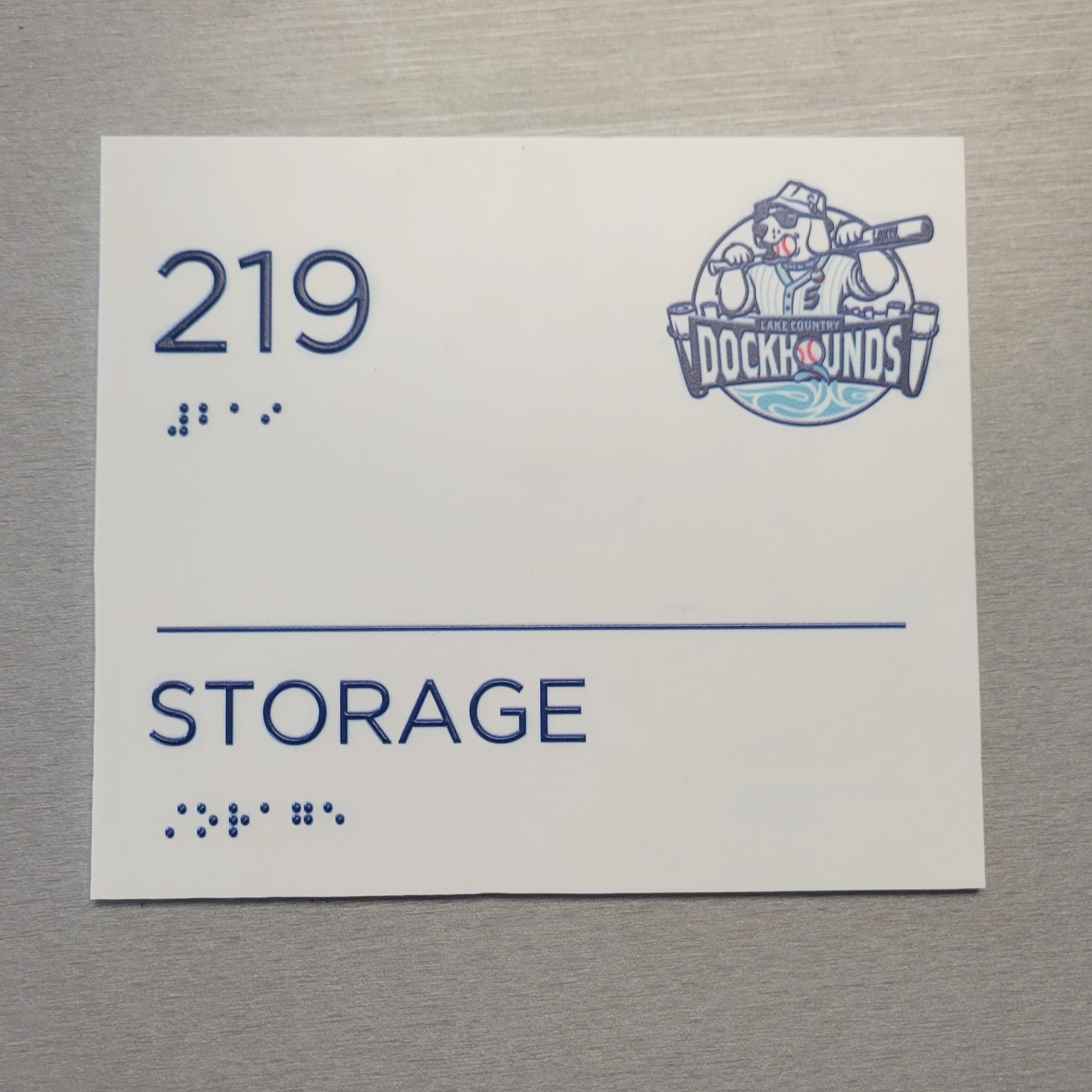 An office plaque with the number 219 and the word "storage" with braille under them. A logo for the Lake Country Dockhounds is featured in the upper right corner.